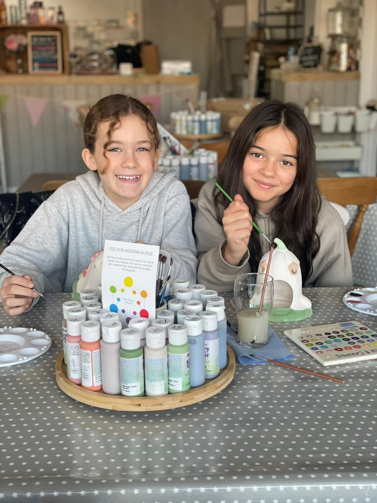 Two children at a table pottery painting with art supplies, including paint bottles and a color chart at Wick & Ceramic, Ickenham, London.