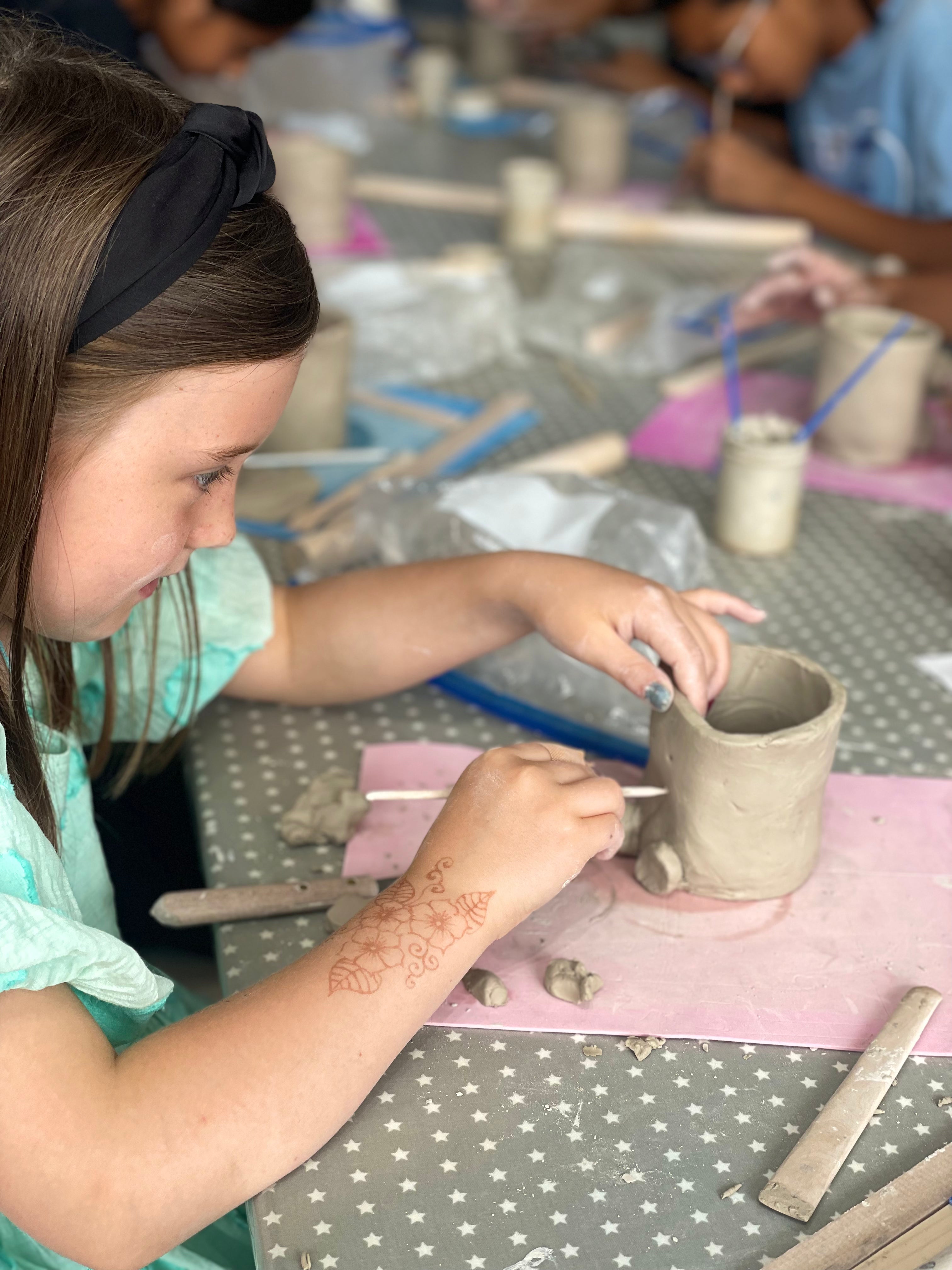 Young girl working on a mug making pottery project from real clay at a table with various pottery items and tools.